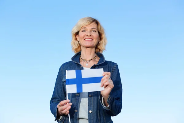 Happy mature woman with flag of Finland standing against the background of a blue sky.