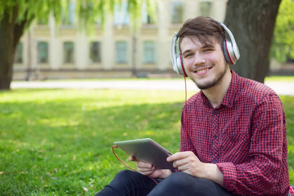 Young man with headphones on his head and a digital tablet in hands sitting on green grass on campus.