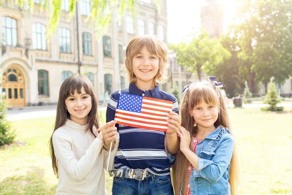 Schoolchildren boy and two girl with the flag of the United States of America in their hands against the background of the college. Training in the USA. Study abroad. School holidays in the US.