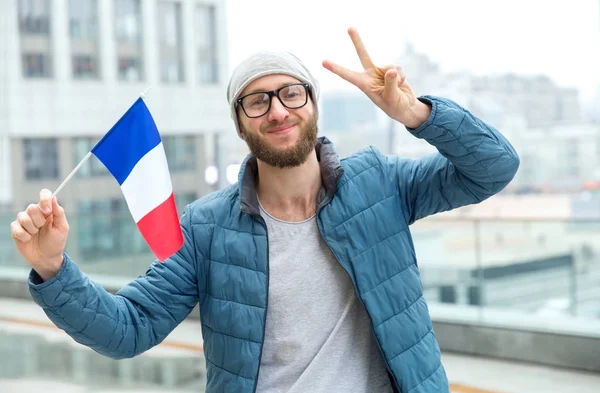 Happy man in glasses with the flag of France shows fingers a sign V, standing on the background of the city. Learn French.