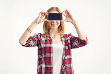 Portre güzel sarışın kadın yüz önünde elinde bir akıllı telefon tutuyor. Boş ekran cep telefonu gösteriliyor. Kız gözleri ışık arka planda ayakta telefon kapattı. Yenilikler teknolojisi.