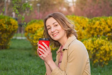 Sabah kahvenizi temiz havada iç. Çiçek açan yeşil bir parkın arka planında elinde bir bardak sıcak çay tutan neşeli bir kadının portresi.