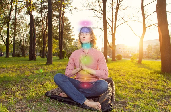 Güzel genç kadın şehir parkında lotus bir poz oturuyor. Kadın gözleri kapalı. Pratik kundalini yoga, meditasyon. Parlayan yedi enerji çakraları. 