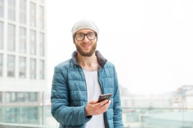 Sakallı ve gözlüklü, elinde akıllı telefon olan adam şehrin arka planında.