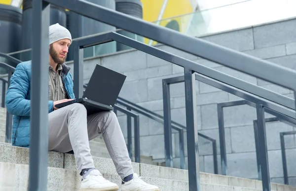 Male freelancer doing remote job using laptop computer. Bearded man working on laptop while sitting  on the steps outdoors at the city.