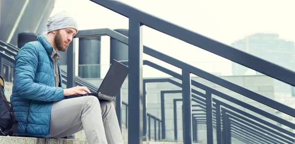 Male freelancer doing remote job using laptop computer. Bearded man working on laptop while sitting  on the steps outdoors at the city.