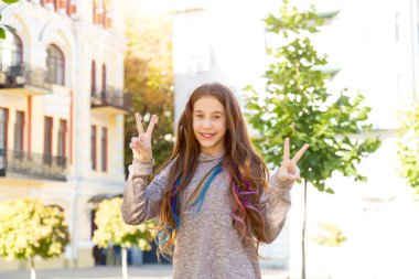 Saçında çok renkli saç örgüleri olan genç bir kız eski bir şehrin arka planında dururken V işareti gösteriyor..