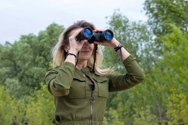 Woman with binoculars looks forward against a background of nature.