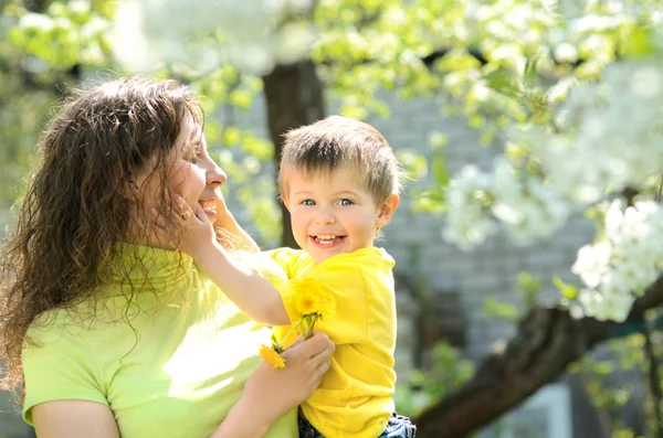 快乐微笑小男孩在他的母亲的胳膊在茂盛的春天庭院苹果 — 图库照片