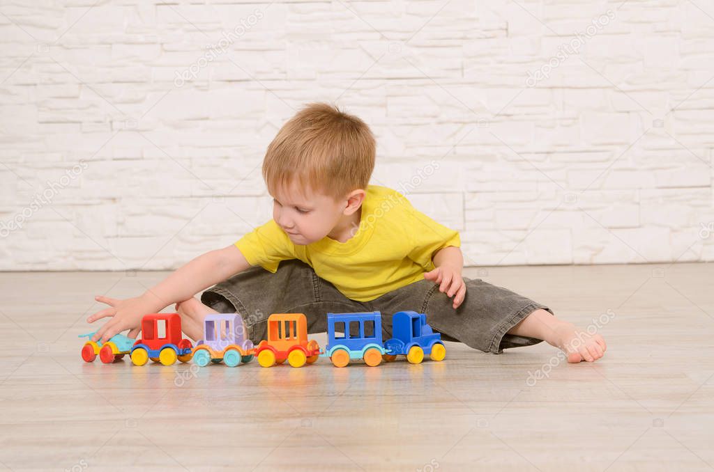 Blond boy sitting on the floor and playing with colored children's train cars against a white brick wall