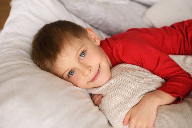 Kırmızı kazaklı yakışıklı çocuk beyaz yastıkların ve battaniyelerin arasında yatıyor.