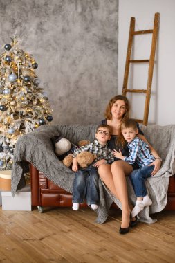 Mutlu ve sevecen bir anne çocuklarını kucaklar. Odadaki koltukta oturur, Noel ağacının arka planına yaslanır. Işıklarla parlar ve bokeh