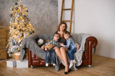 Mutlu ve sevecen bir anne çocuklarını kucaklar. Odadaki koltukta oturur, Noel ağacının arka planına yaslanır. Işıklarla parlar ve bokeh
