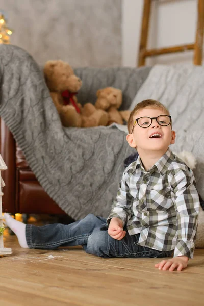 戴眼镜的男孩坐在木制的一半沙发上 沙发上铺着毛织的灰色格子布 上面挂着玩具熊 — 图库照片
