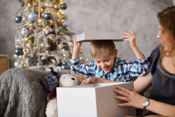 Boy was delighted and surprised at gift he saw in big gray box, in his mothers hands, sitting on sofa against background of Christmas tree glowing with them bokeh