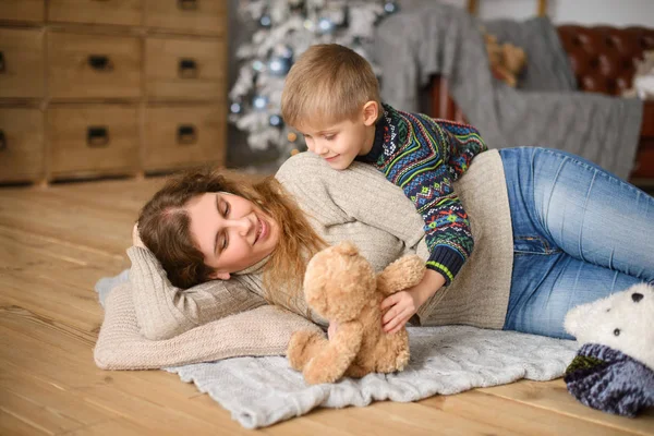 Mom and son playing with teddy bear toy lying on  wooden floor, against background of interior, with Christmas tree with lights, bokeh