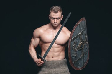 Kılıç ve kalkan siyah arka plan üzerine izole poz vücut geliştirmeci adam. Ciddi gömleksiz adam mascular vücudunu gösteren