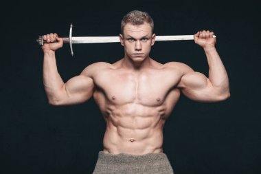 Vücut geliştirmeci adam siyah arka plan üzerine izole bir kılıç ile poz. Ciddi gömleksiz adam mascular vücudunu gösteren