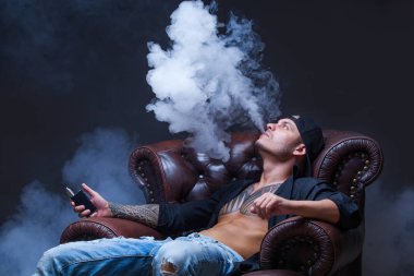 Vaper. Dövmeli adam koyu arka plan üzerinde elektronik sigara duman deri kanepede oturuyor.