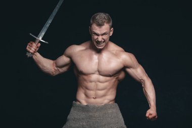 Vücut geliştirmeci adam siyah arka plan üzerine izole bir kılıç ile poz. Ciddi gömleksiz adam mascular vücudunu gösteren