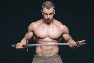 Vücut geliştirmeci adam siyah arka plan üzerine izole bir kılıç ile poz. Ciddi gömleksiz adam mascular vücudunu gösteren