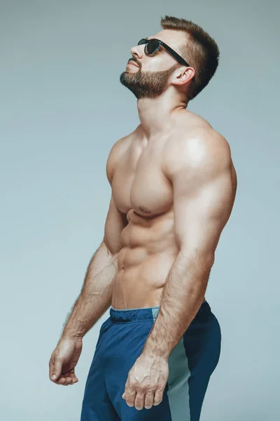 .fisiculturista posando. Bonito desportista poder masculino. Fitness musculado em calções azuis e óculos de sol. sobre fundo cinzento isolado. Homem com tronco musculado. Torso forte do modelo da aptidão do homem atlético — Fotografia de Stock