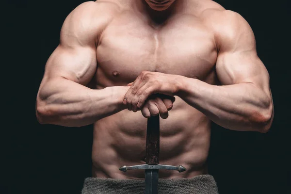 Bodybuilder man posing with a sword isolated on black background. Serious shirtless man demonstrating his mascular body. hands on a sword — Stock Photo, Image