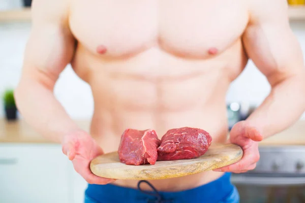 Homem musculoso com um tronco nu na cozinha com um pedaço de carne bovina, o conceito de uma dieta saudável. Modo de vida atlético . — Fotografia de Stock