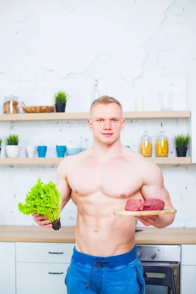 Homem musculoso com um tronco nu na cozinha com uma salada e um pedaço de carne bovina, o conceito de uma dieta saudável. Modo de vida atlético . — Fotografia de Stock