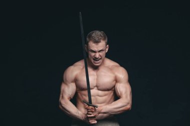 Vücut geliştirmeci adam siyah arka plan üzerine izole bir kılıç ile poz. Ciddi gömleksiz adam mascular vücudunu gösteren