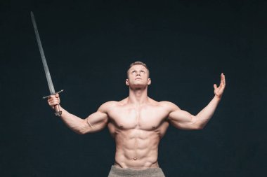 Vücut geliştirmeci adam siyah arka plan üzerine izole bir kılıç ile poz. Ciddi gömleksiz adam mascular vücudunu gösteren