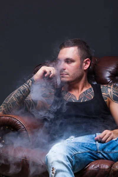 Vaper. Mannen med tatueringar som sitter på en skinnsoffa som rök en elektronisk cigarett på den mörka bakgrunden. — Stockfoto