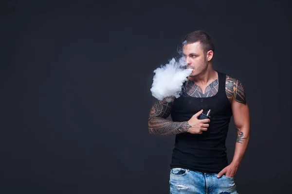 Vaper. Mannen med en muskulös överkropp med tatueringar röka en elektronisk cigarett på den mörka bakgrunden — Stockfoto