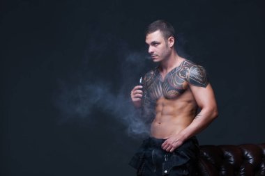 Vaper. Dövmeli bir kaslı çıplak gövde adamla koyu arka plan üzerinde elektronik sigara duman