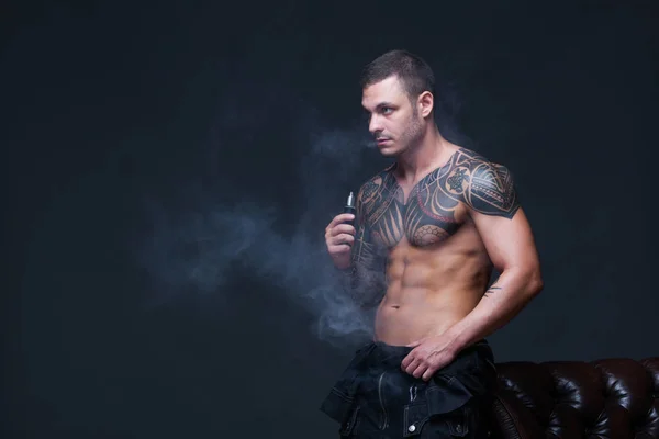 Vaper. El hombre con un torso desnudo muscular con tatuajes fuma un cigarrillo electrónico en el fondo oscuro —  Fotos de Stock