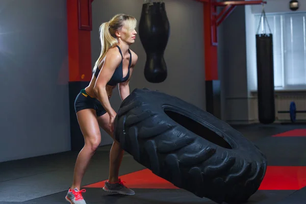 Apto atleta feminino exercitar-se com um pneu enorme, girando e transportar no ginásio. Crossfit mulher exercitando com pneu grande — Fotografia de Stock