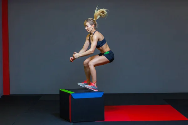 Atleta de fitness feminino bonita executa saltos caixa em um ginásio escuro vestindo top de esportes preto e meia-calça curta com o rosto escondido . — Fotografia de Stock