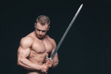 Vücut geliştirmeci adam siyah arka plan üzerine izole bir kılıç ile poz. Ciddi gömleksiz adam mascular vücudunu gösteren