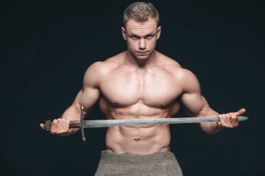 Vücut geliştirmeci adam siyah arka plan üzerine izole bir kılıç ile poz. Ciddi gömleksiz adam mascular vücudunu gösteren