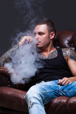 Vaper. Dövmeli adam koyu arka plan üzerinde elektronik sigara duman deri kanepede oturuyor.