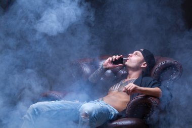 Vaper. Dövmeli adam koyu arka plan üzerinde elektronik sigara duman deri kanepede oturuyor.