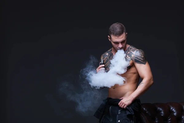 Vaper. El hombre con un torso desnudo muscular con tatuajes fuma un cigarrillo electrónico en el fondo oscuro — Foto de Stock