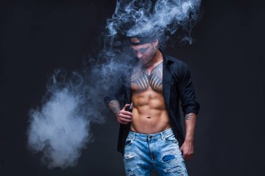 Vaper. Adam mavi kot pantolon giymiş, siyah gömlek ve siyah beyzbol şapkası dövmeler ile koyu arka plan üzerinde elektronik sigara duman