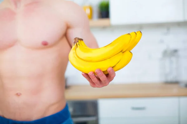 Homem musculoso com um tronco nu na cozinha com frutas, conceito de alimentação saudável. Estilo de vida atlético. Bananas close-up . — Fotografia de Stock