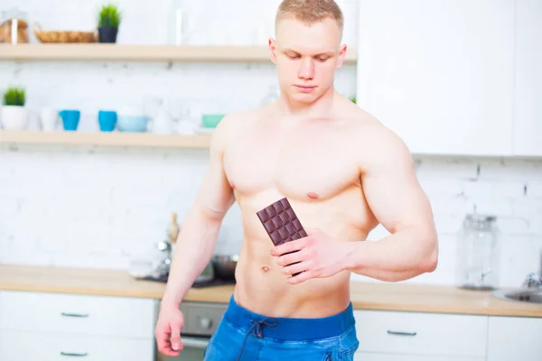 Homem muscular com um tronco nu na cozinha com uma telha de chocolate, o conceito de uma dieta saudável. Estilo de vida atlético. A tentação de doce . — Fotografia de Stock