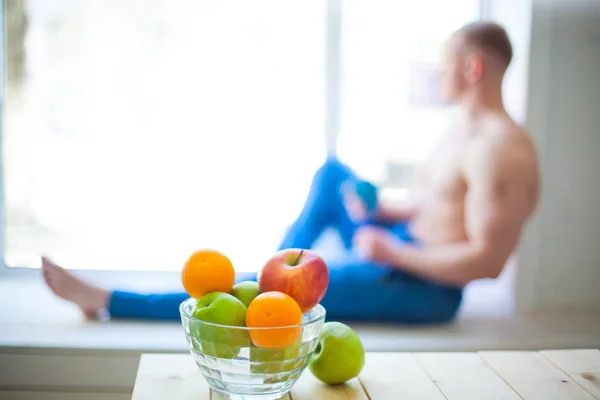 Mesa com frutas, e no fundo está fora de foco jovem homem bonito sentado com peito nu na janela com xícara de chá — Fotografia de Stock