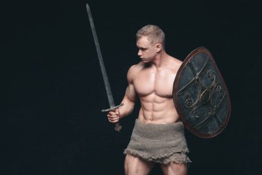 Kılıç ve kalkan siyah arka plan üzerine izole poz vücut geliştirmeci adam. Ciddi gömleksiz adam mascular vücudunu gösteren
