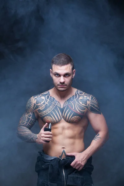 Vaper. El hombre con un torso desnudo muscular con tatuajes fuma un cigarrillo electrónico en el fondo oscuro — Foto de Stock