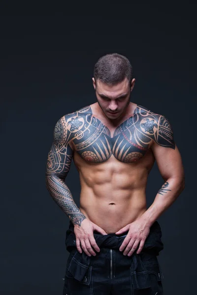 Der Mann mit dem muskulösen nackten Oberkörper mit Tätowierungen auf dem dunklen Hintergrund. abs — Stockfoto