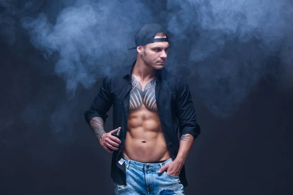 汽.身着蓝色牛仔裤, 黑色衬衫和黑色棒球帽的男子纹身烟雾在黑暗的背景上的电子香烟 — 图库照片